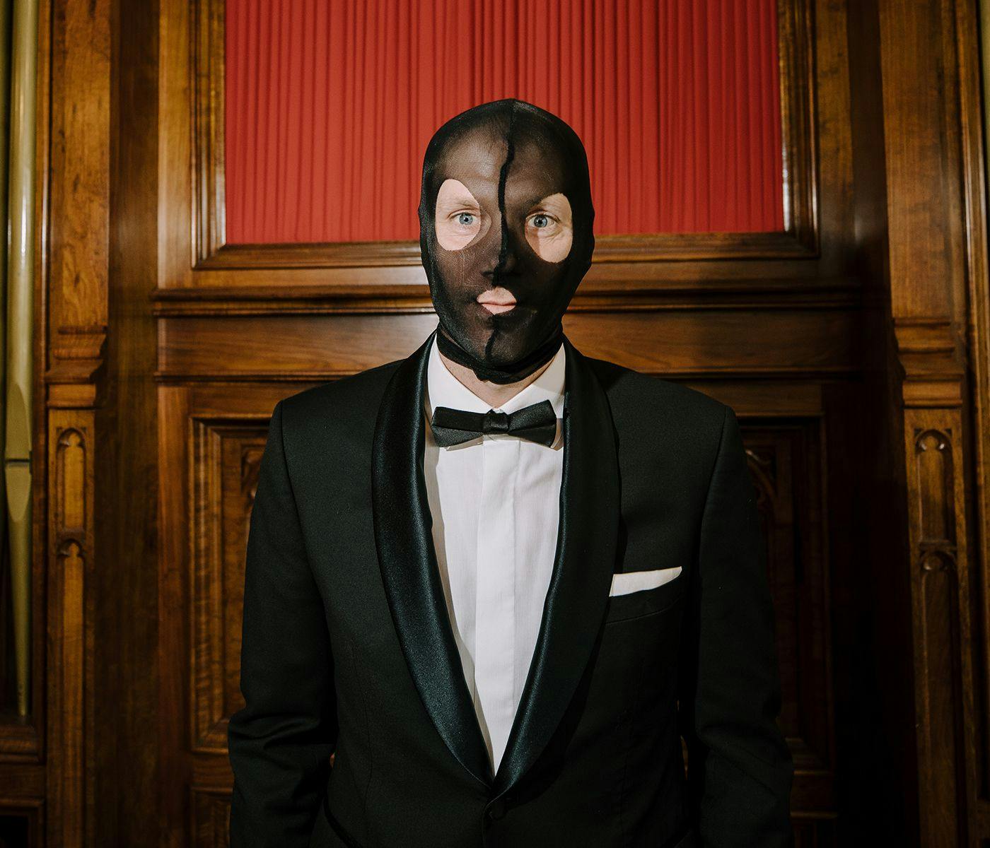 A person wearing a black tuxedo and bow tie. Their face is covered with a ski-mask style stocking.