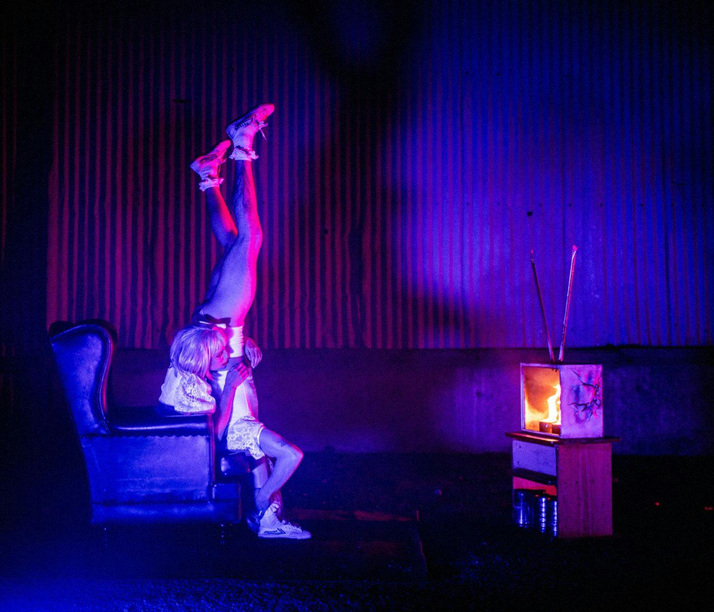 Two people sit on a chair watching a burning TV. One person is upside down with their legs in the air.