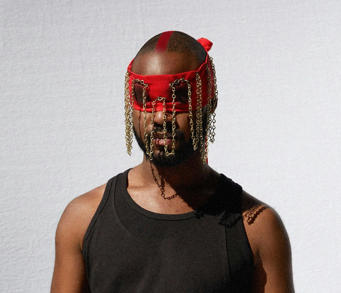 Genesis Owusu wearing a black tank-top and a ornate red chained bandana, covering their eyes.